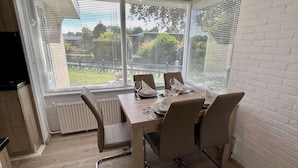 Ein Essbereich mit Tisch für vier Personen, großen Fenstern mit Jalousien und einer weißen Ziegelwand.
