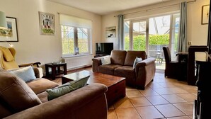 Ein Wohnzimmer mit braunen Sofas, einem Fernseher und einem Klavier. Es gibt Fenster mit Vorhängen, eine Heizung und ein gerahmtes Bild an der Wand.