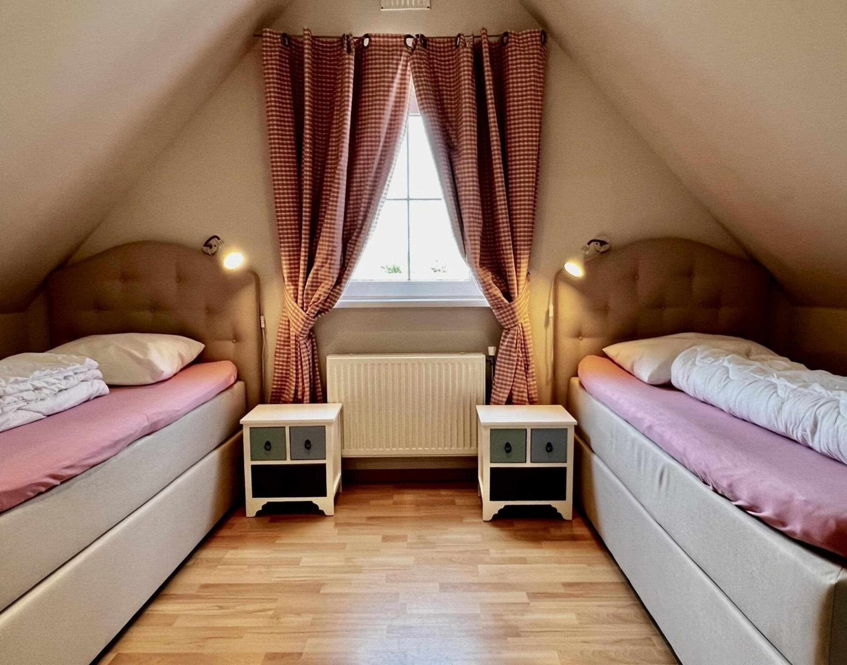 A small, cozy attic bedroom with two beds, a window with checkered curtains, and a radiator.