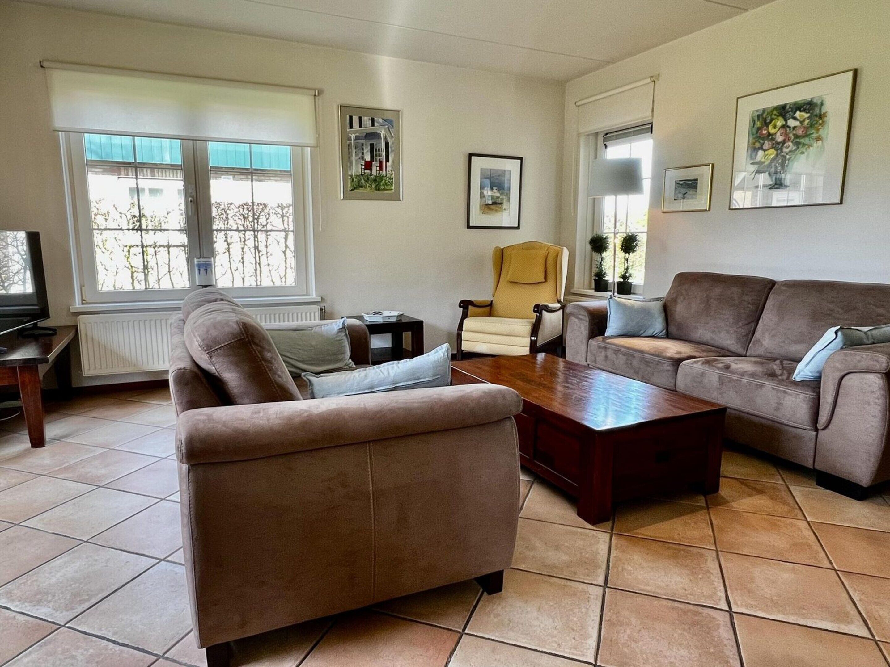 A living room with two sofas, a coffee table, a TV, and a chair. There are windows with blinds, a radiator, and framed pictures on the walls.