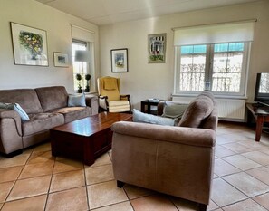 A living room with a brown sofa, a yellow armchair, and a coffee table. There are framed pictures on the walls and a window with a view of trees. - Holiday Home in Sint Maartenszee for 6 (Sint Maartensvlotbrug)