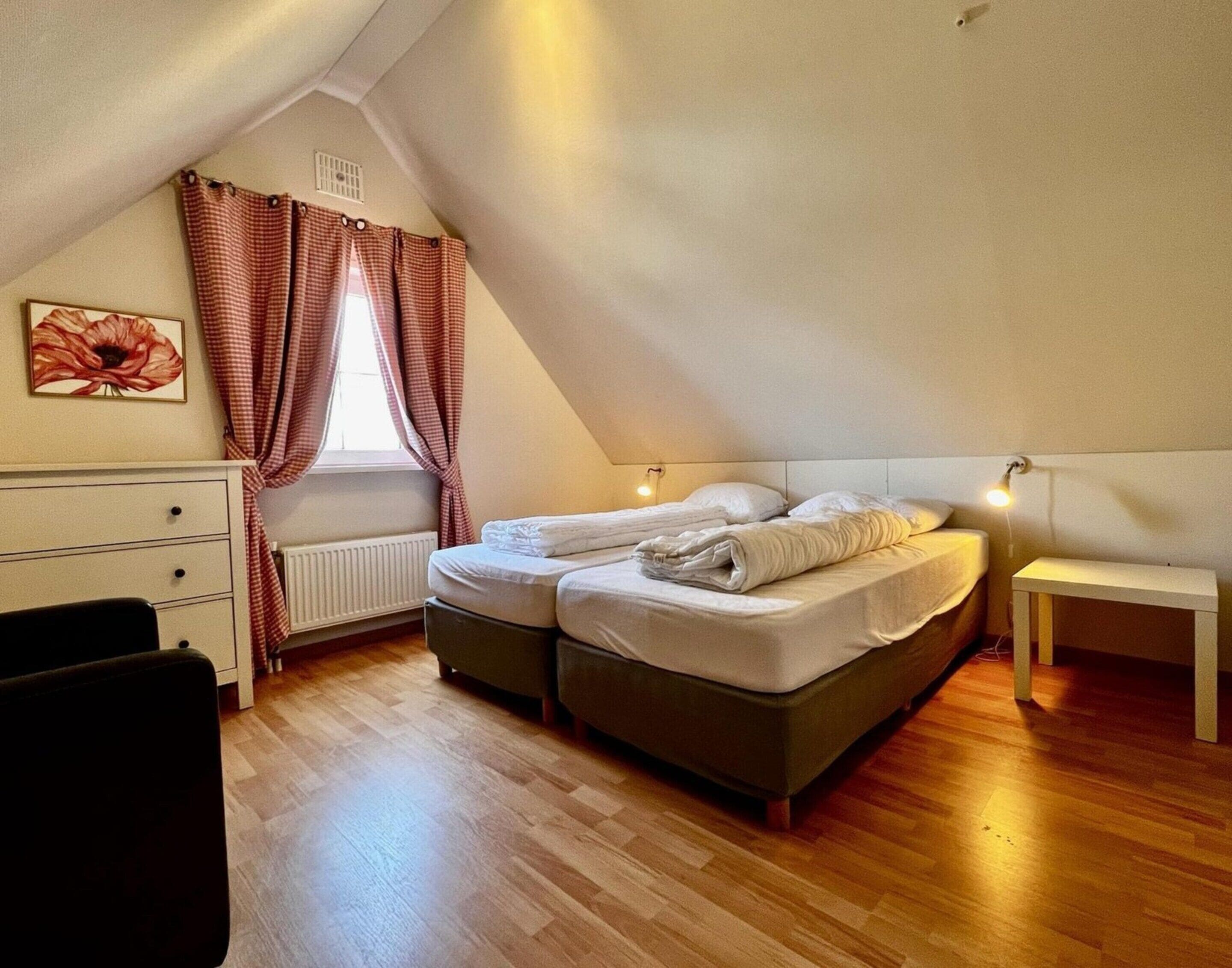 Une chambre mansardée chaleureuse, avec un plafond en pente, un lit avec deux oreillers, une table de chevet, un radiateur et une fenêtre ornée de rideaux à carreaux.