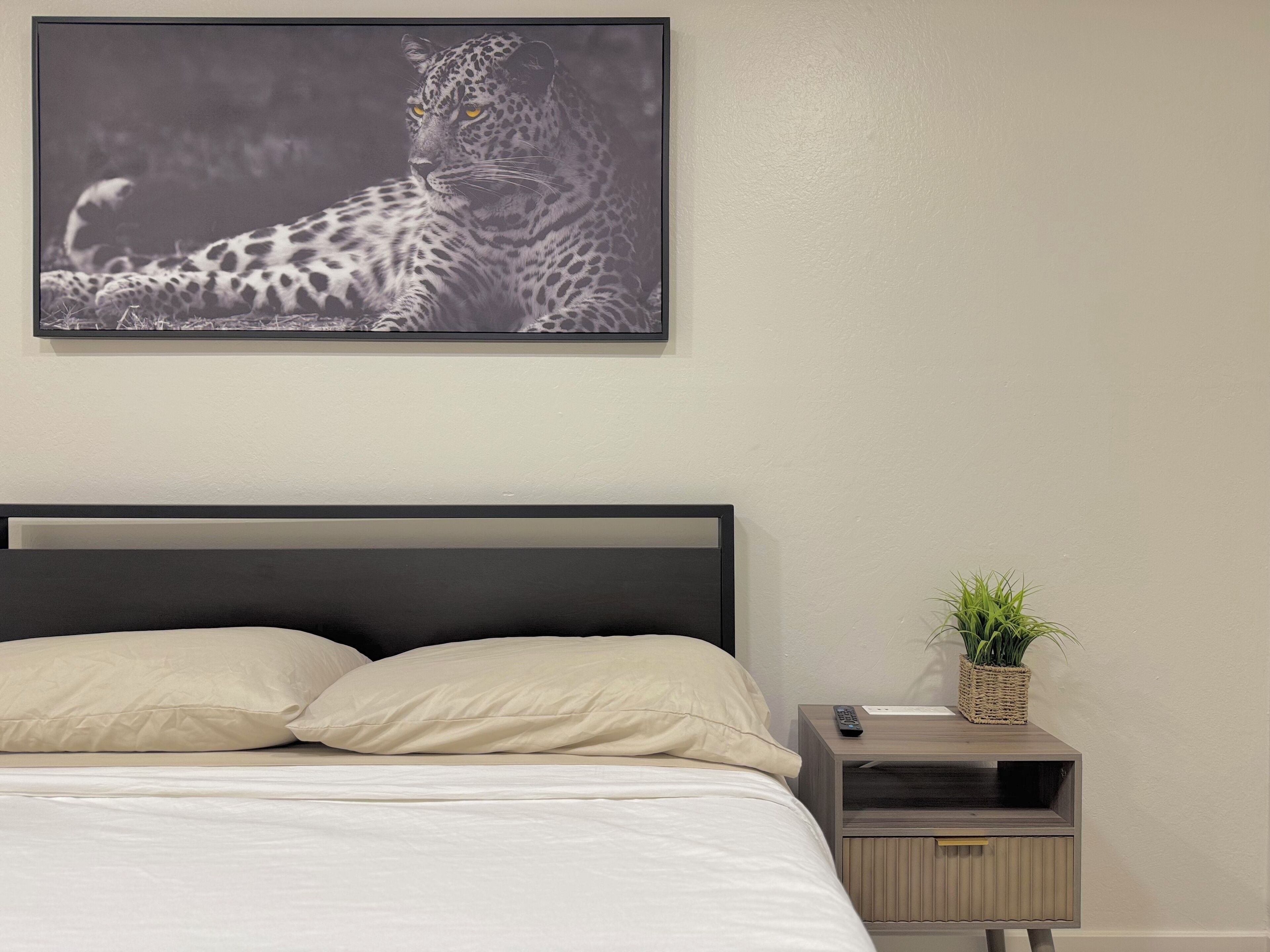 Une chambre à coucher avec un lit, des oreillers, une table de chevet avec une plante et un tableau encadré au mur.