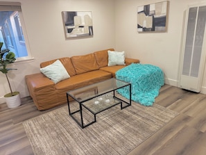 A living room with a brown leather sofa, two white pillows, a glass coffee table, a blue throw blanket, and two abstract paintings on the wall.