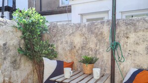 A small outdoor seating area with a wooden table, two chairs, a potted plant, and a hanging green rope.
