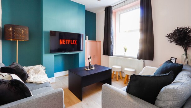 A modern living room with teal walls, a black coffee table, grey sofas, and a TV displaying 'NETFLIX'.