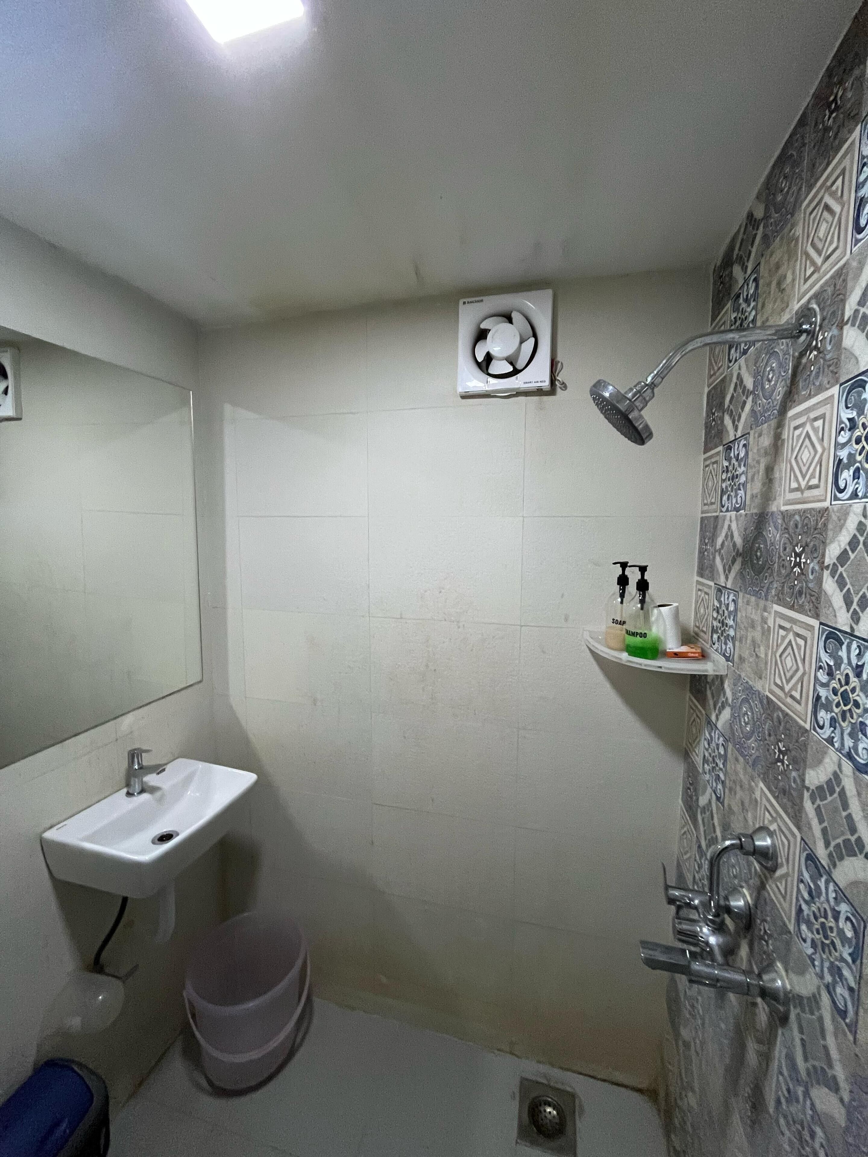 Casa de banho pequena com lavatório branco, chuveiro manual, sanita e prateleira com artigos de higiene pessoal.