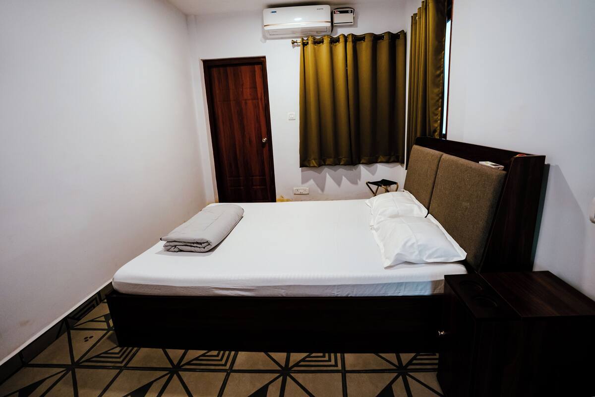 A hotel room with a bed, a bedside table, a wall-mounted air conditioner, a window with curtains, and a door.