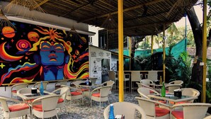 An outdoor seating area with wicker chairs and tables, a mural with cosmic and abstract designs, and a green canopy overhead. - Half Orange (Vagator)