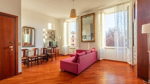 A living room with a wooden floor, a pink sofa, a dining table with chairs, and a bookshelf.