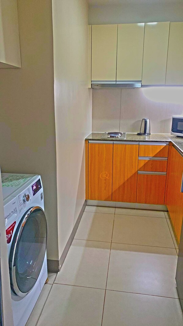 A compact kitchen with a washing machine, wooden cabinets, and a tiled floor. - One Manchester Place 1BR Ocean View (Lapu-Lapu)