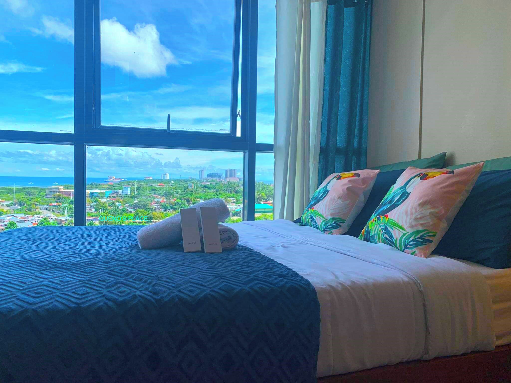 A hotel room with a large window offering a cityscape view, a neatly made bed with patterned bedding, and tropical-themed pillows.