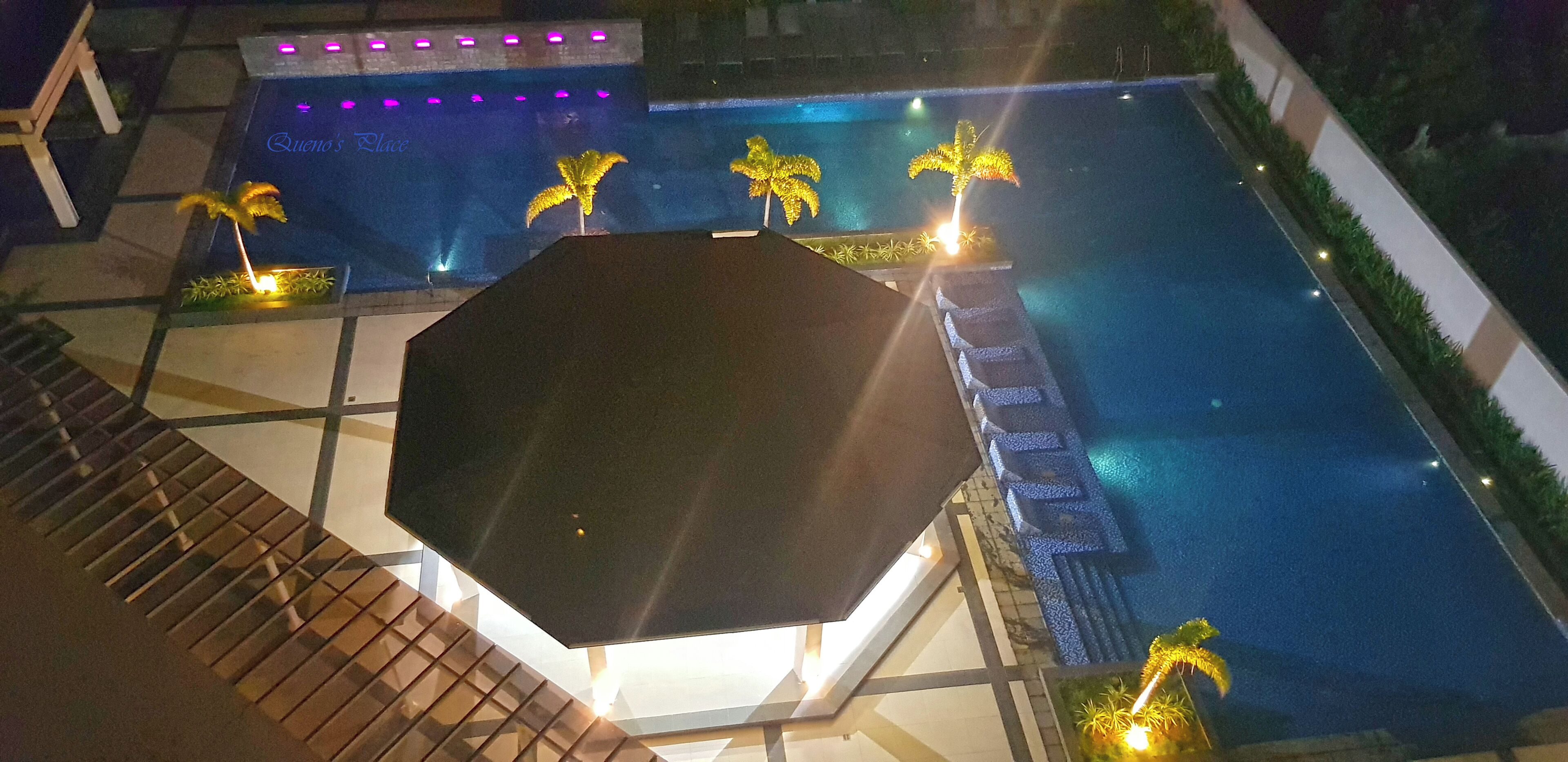 Un área de piscina en la azotea con techo de vidrio geométrico, palmeras y un letrero que dice "Queno's Place".