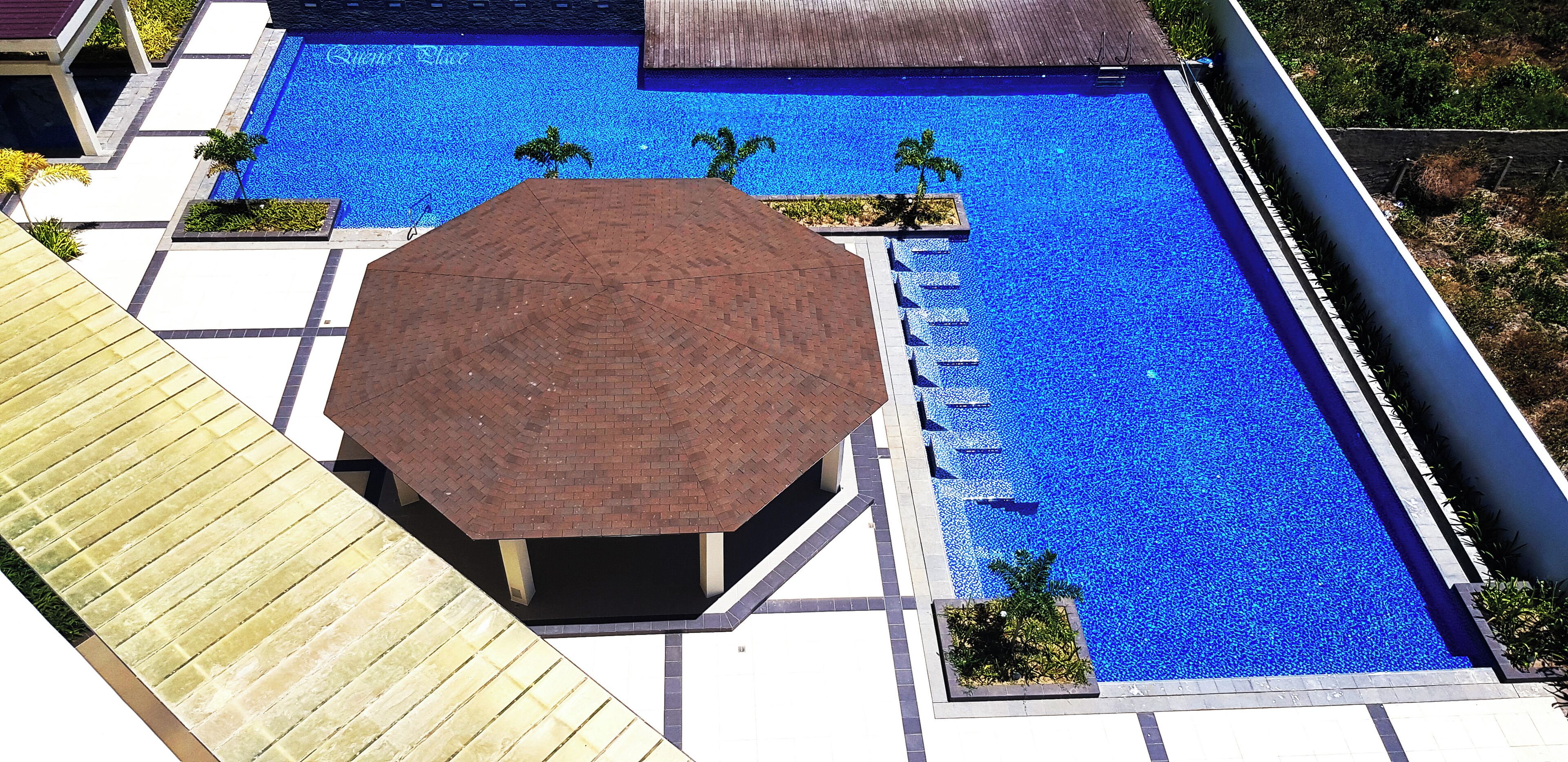 A large rectangular swimming pool with a tiled deck surrounding it, a covered seating area, and small palm trees.