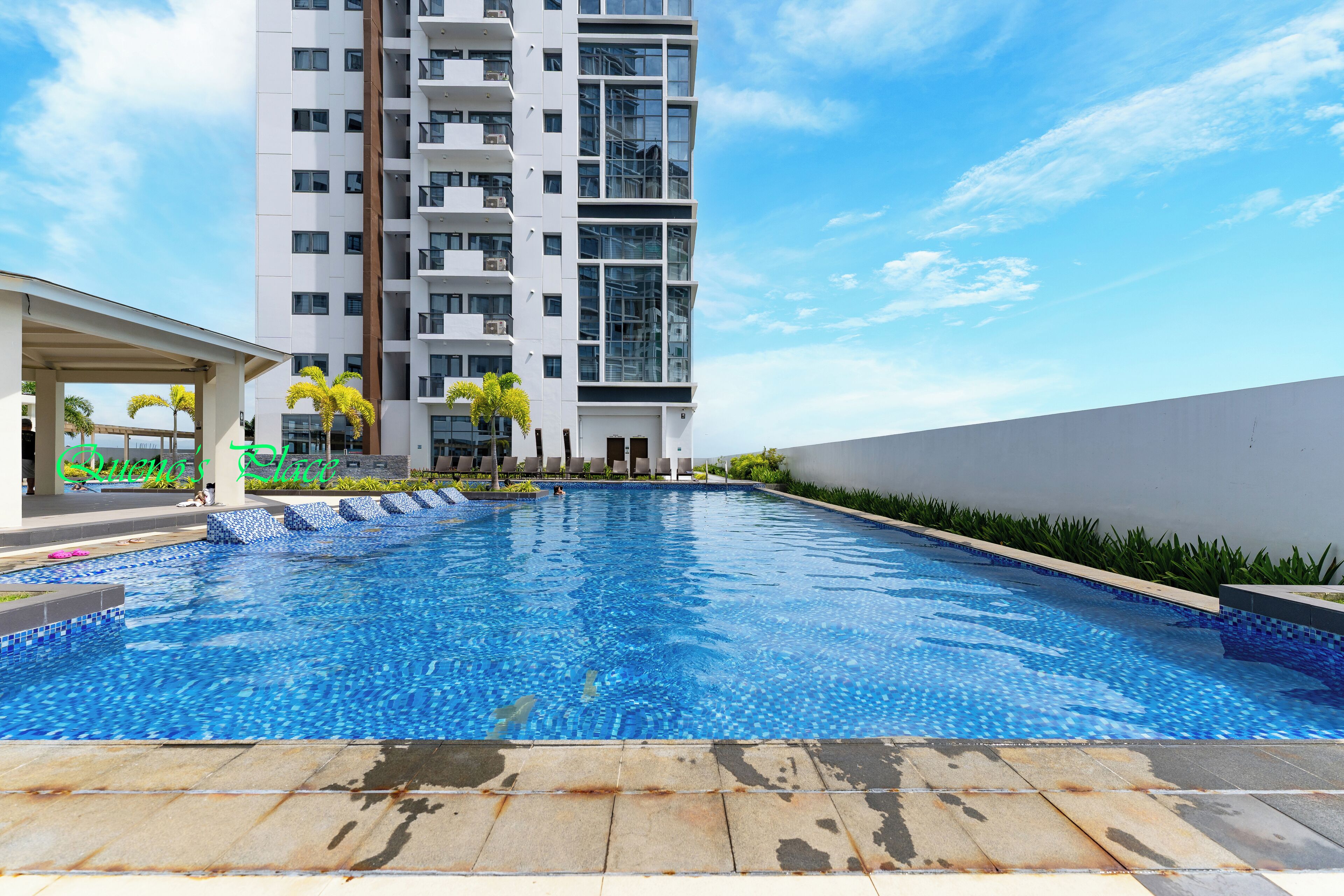 A swimming pool with a tiled deck, surrounded by a high-rise building and a covered area with palm trees.