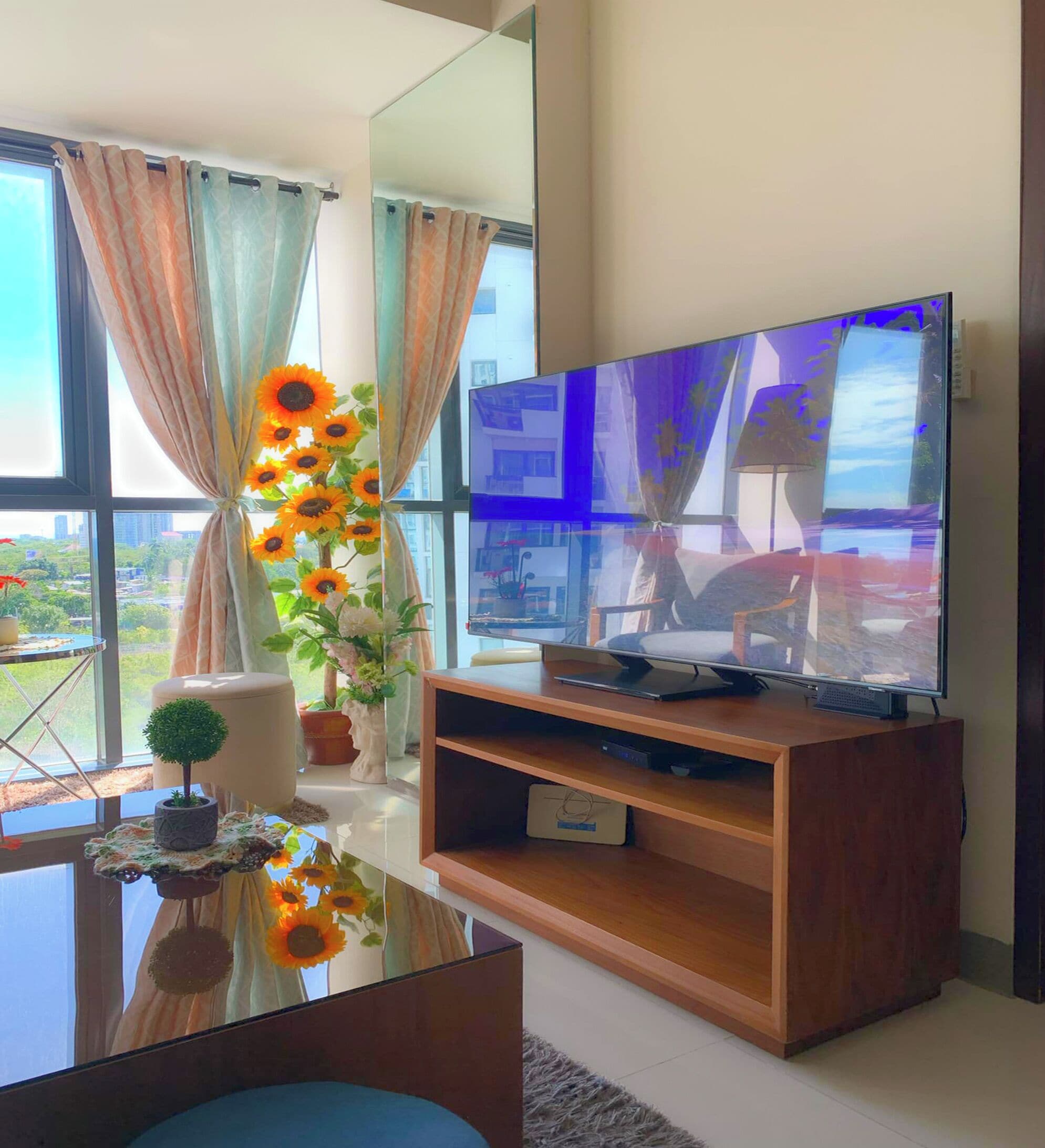 A modern living room with a flat-screen TV on a wooden stand, a glass coffee table, and a view of a cityscape through large windows.