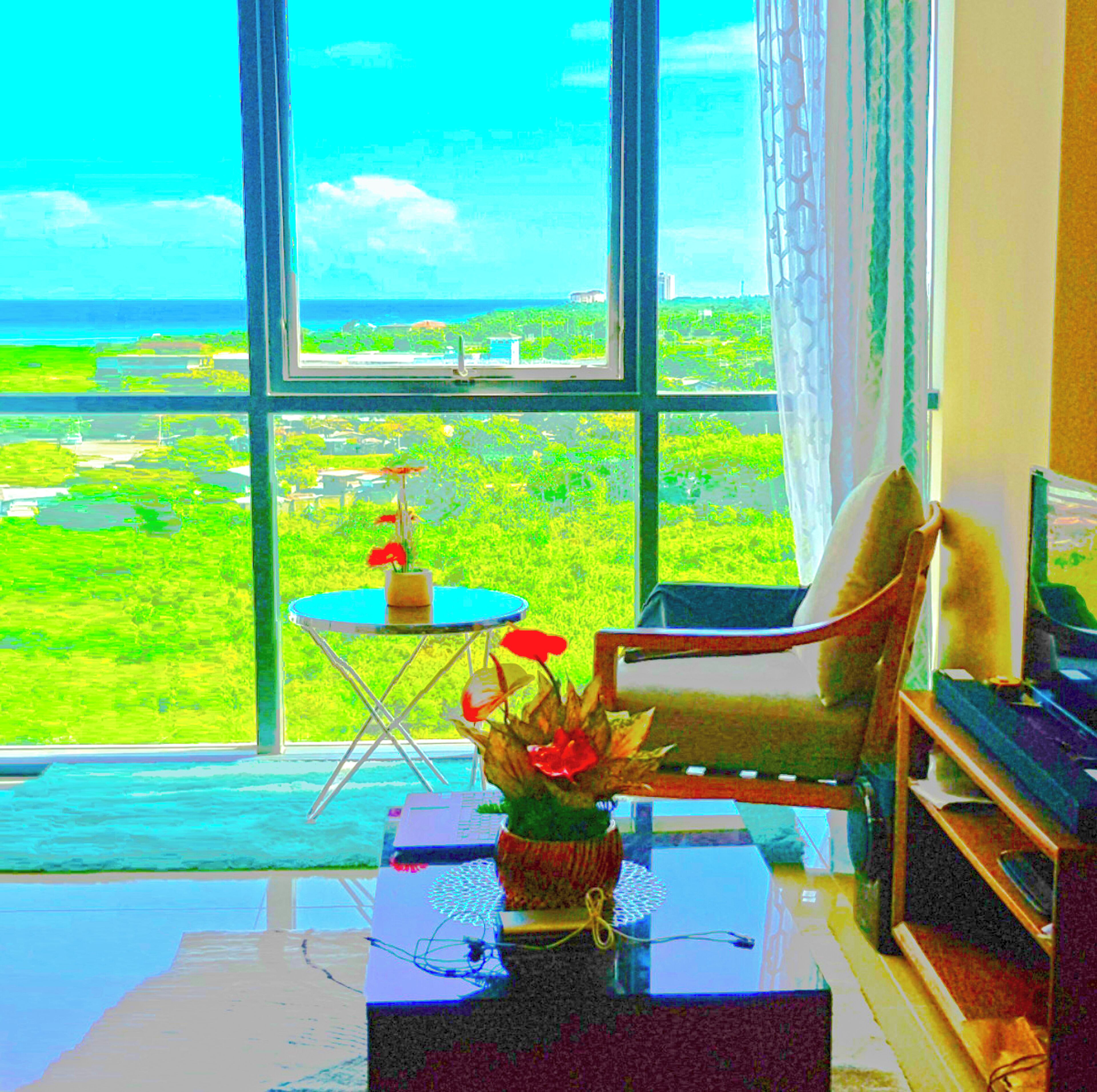 A room with a large window offering a view of a green landscape and the ocean. There is a chair and a small table with a vase of flowers.