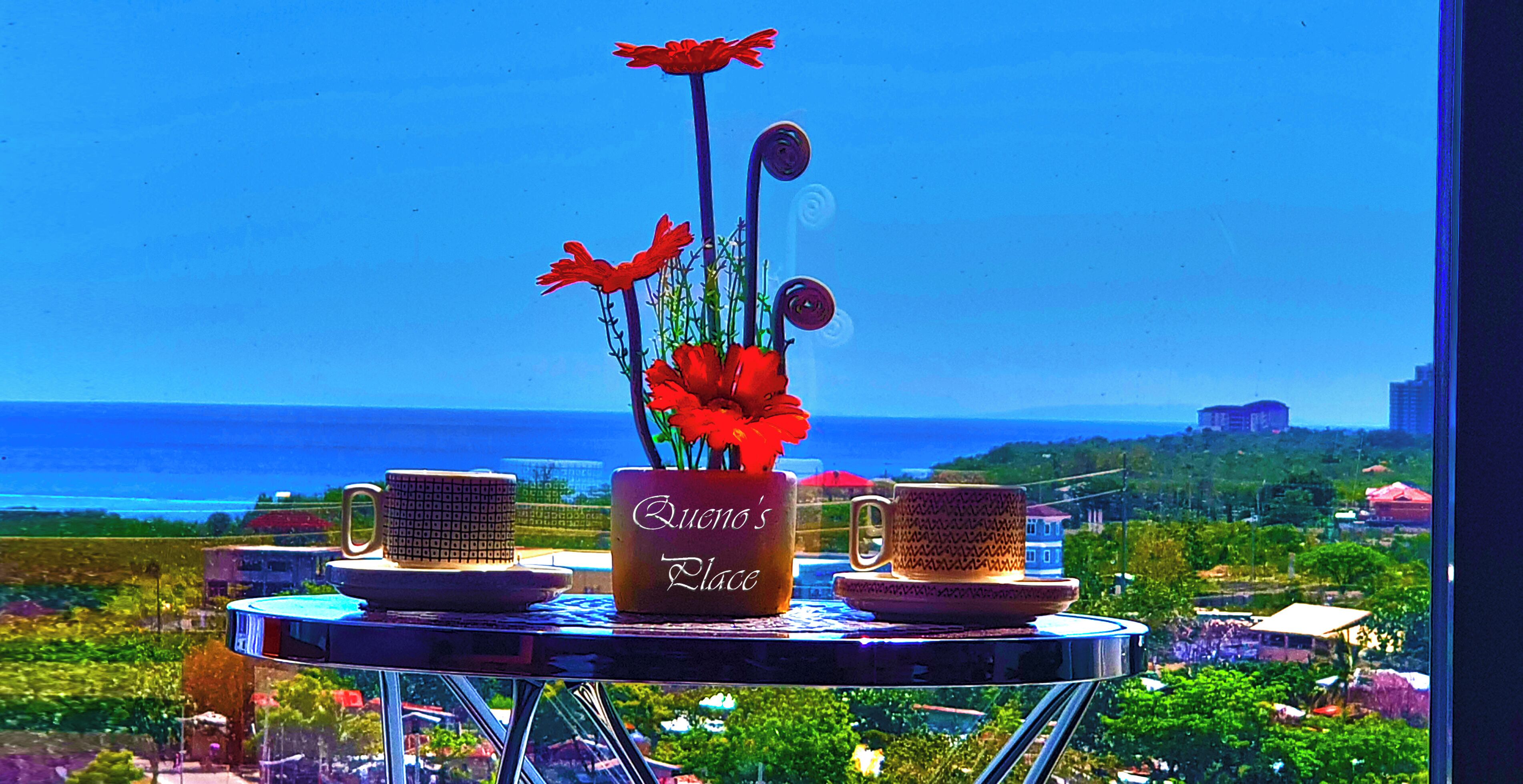 Una mesa con un jarrón de flores rojas y dos tazas decoradas, con vista al mar.