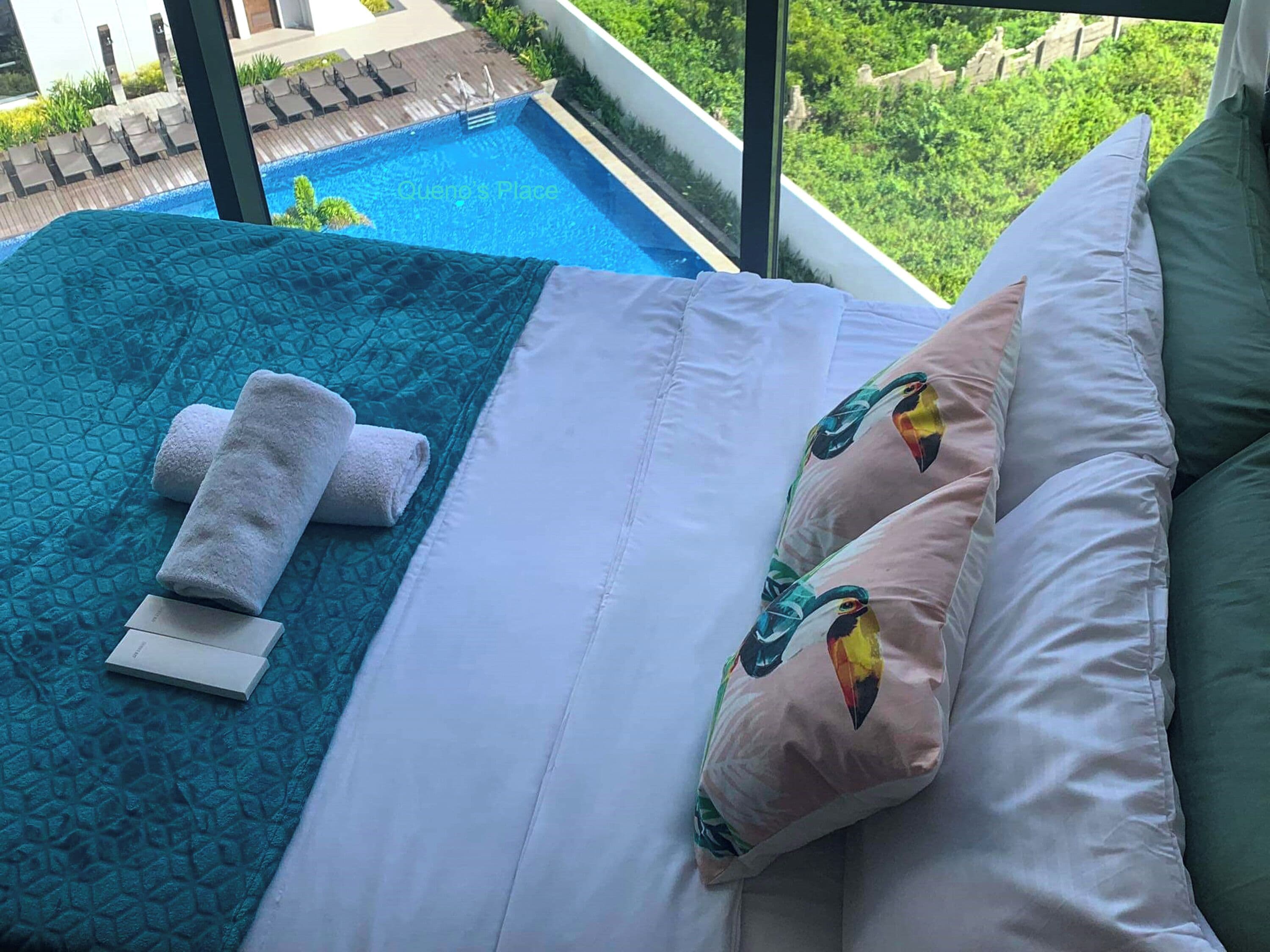 A hotel room with a bed, a blue textured blanket, white pillows, and a view of a swimming pool and greenery.