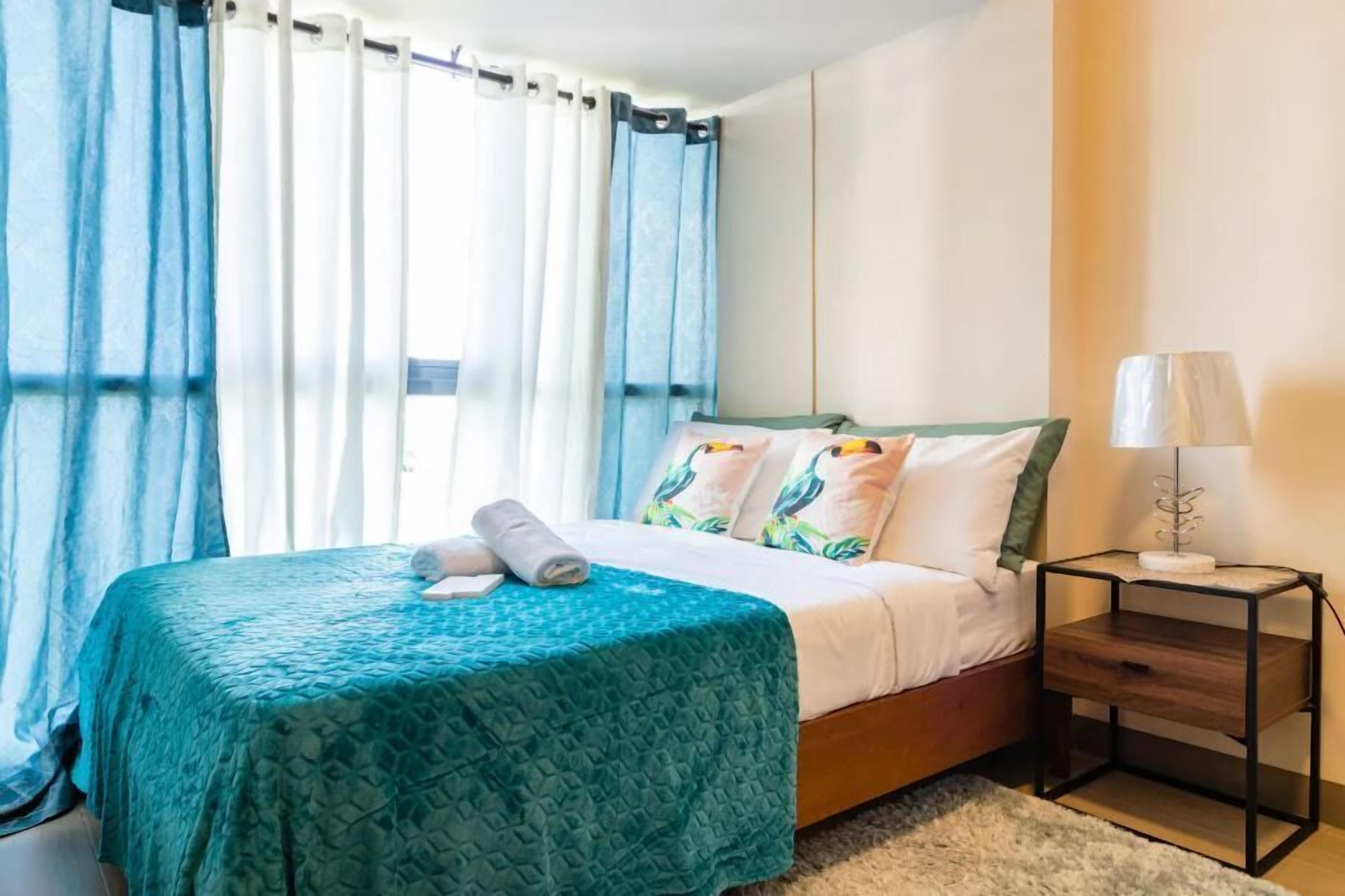 Una cama bien hecha con una colcha verde azulada, un cojín con un diseño de pájaro y una lámpara de noche sobre una mesita de madera.