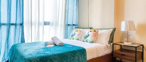 Una cama bien hecha con una colcha verde azulada, un cojín con un diseño de pájaro y una lámpara de noche sobre una mesita de madera.
