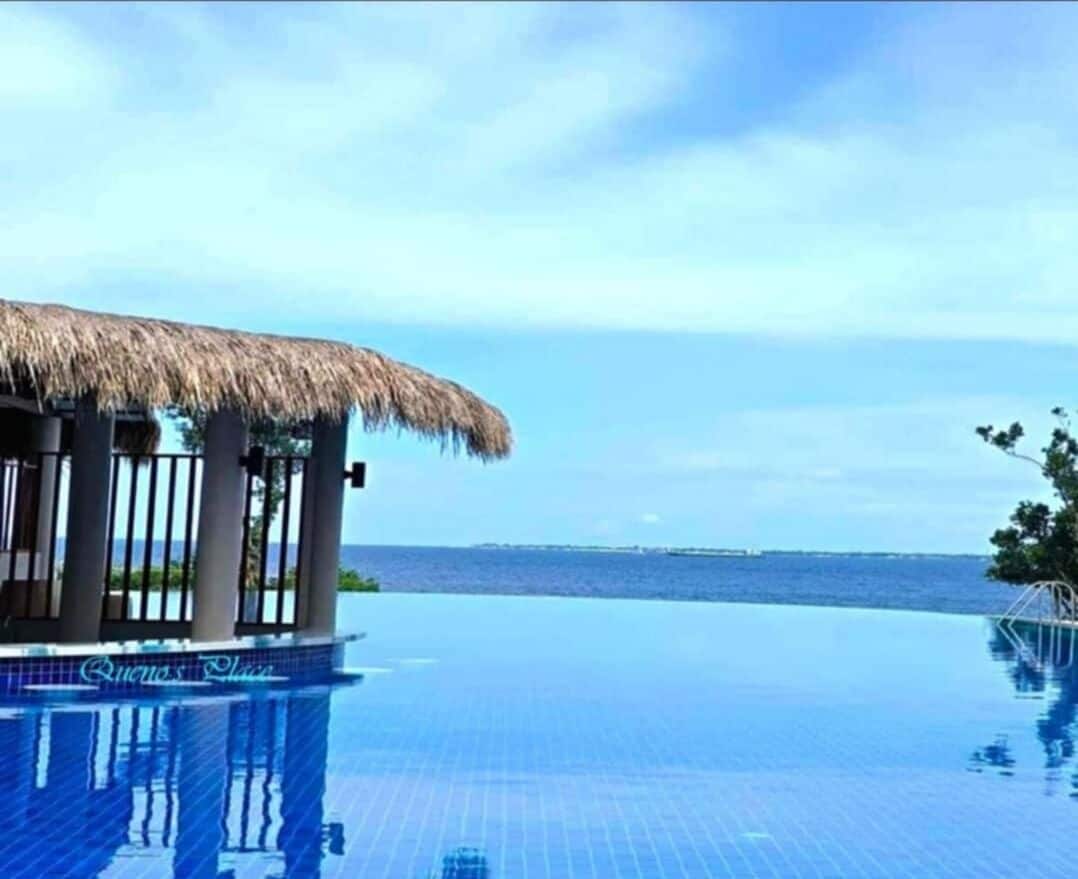 Una piscina infinita con una estructura de techo de paja, vista despejada al océano y el texto 'Queena Place' en el borde de la piscina.