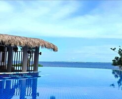 Una piscina infinita con una estructura de techo de paja, vista despejada al océano y el texto 'Queena Place' en el borde de la piscina.