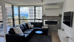 Ein modernes Wohnzimmer mit einer schwarzen Ledersofa, einem kleinen runden Tisch und einem großen Fenster mit Blick auf das Meer und die Gebäude.