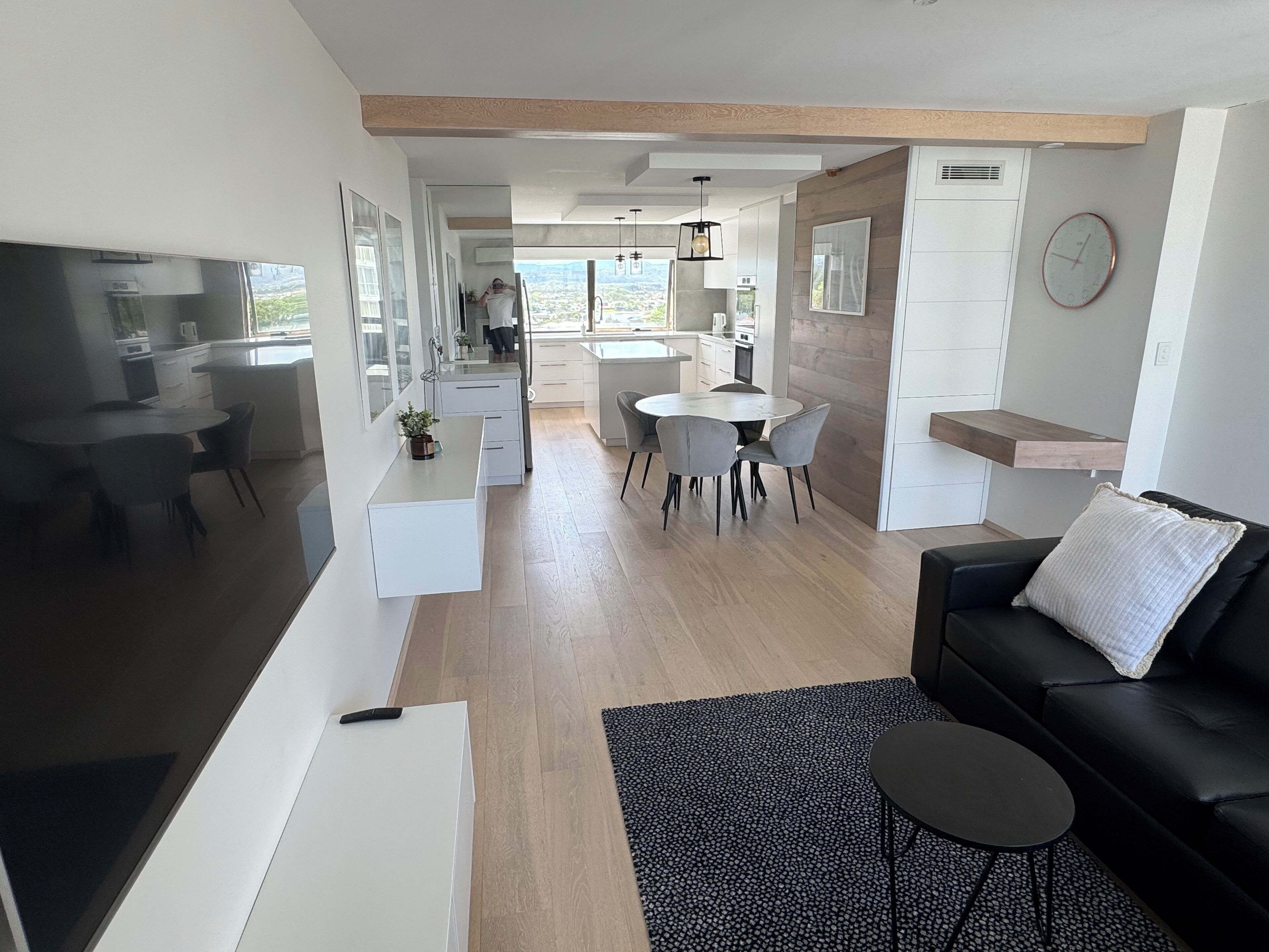 Uma sala de estar moderna com um sofá de couro preto, uma mesa pequena preta e uma área de jantar com uma mesa redonda e cadeiras cinzas.