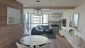 Ein modernes Wohnzimmer mit einer Couch, einem Esstisch mit Stühlen, einem Kamin und einem großen Fenster mit Blick auf Gebäude.
