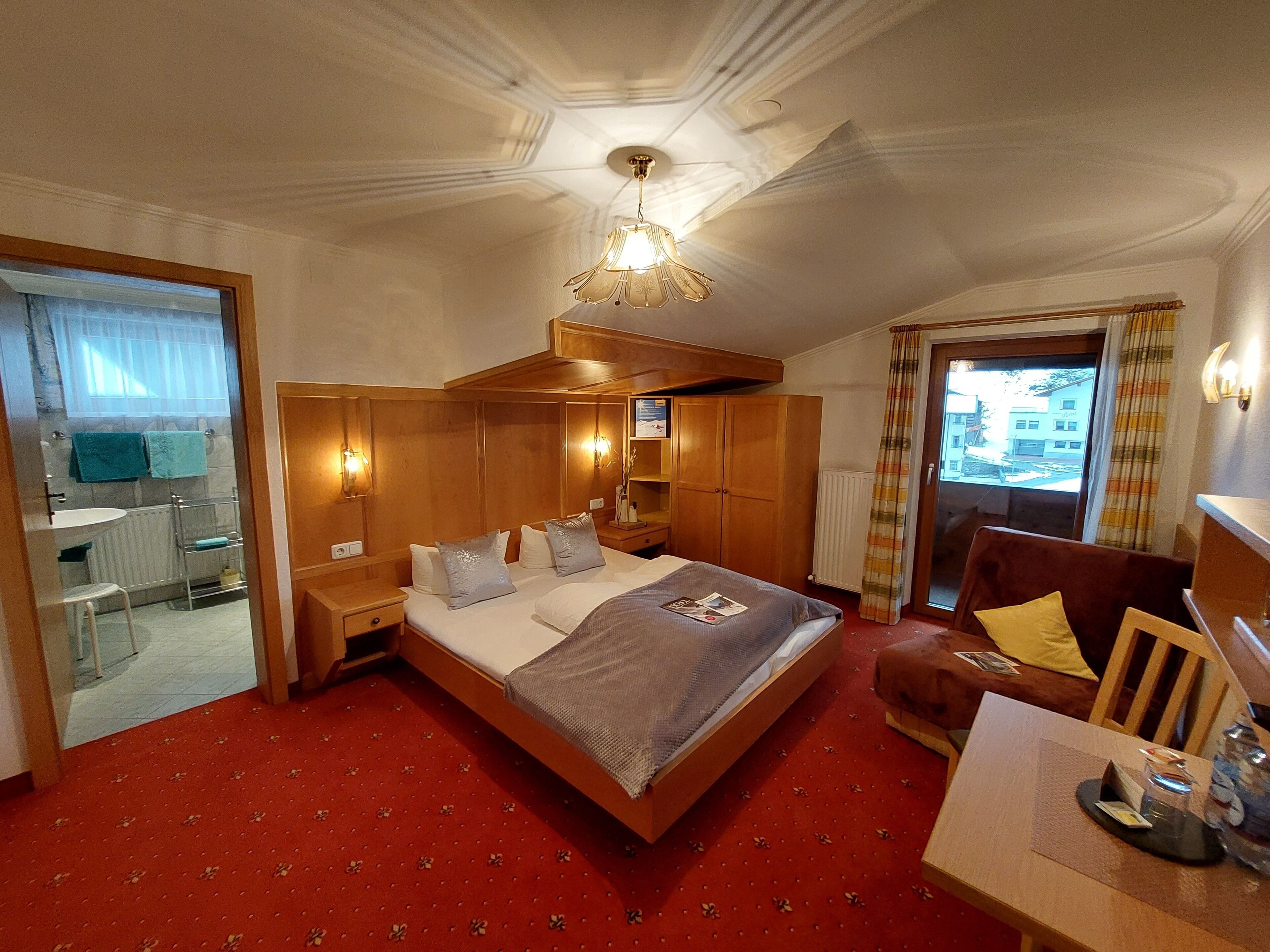 A hotel room with a large bed, wooden headboard, a small table with a lamp, a window with curtains, and a view of buildings outside.