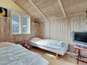 A room with wooden walls and ceiling, a bed with a white mattress and light blue bedding, a television on a stand, and a window with blinds.