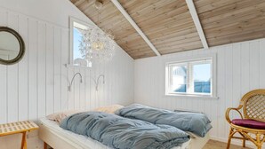 Ein gemütliches Schlafzimmer mit Holzboden, einem Bett mit blauer Bettwäsche, einem Korbsessel und einer weißen Wand mit Spiegel und Wandleuchten.