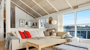 Ein großzügiges Wohnzimmer mit hohen Decken, Holzbalken, einem großen Fenster mit Aussicht, einer Sofagarnitur, einem Couchtisch und einem Bücherregal.