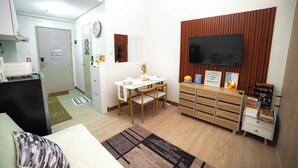 A compact living space with a dining area, a TV mounted on a wooden panel, a small cabinet with drawers, and a visible door leading to another room.