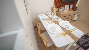 A dining area with a long white table set with plates, bowls, and cutlery. There are wooden chairs around the table, and a small cabinet with decorative items is in the background.
