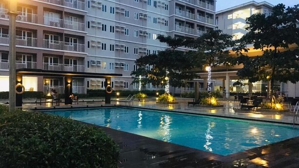 A swimming pool with surrounding lounge areas, a multi-story building with balconies, and outdoor lighting.