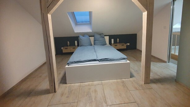 A bedroom with a sloped ceiling, a skylight, a bed, bedside tables, and a view through a window.