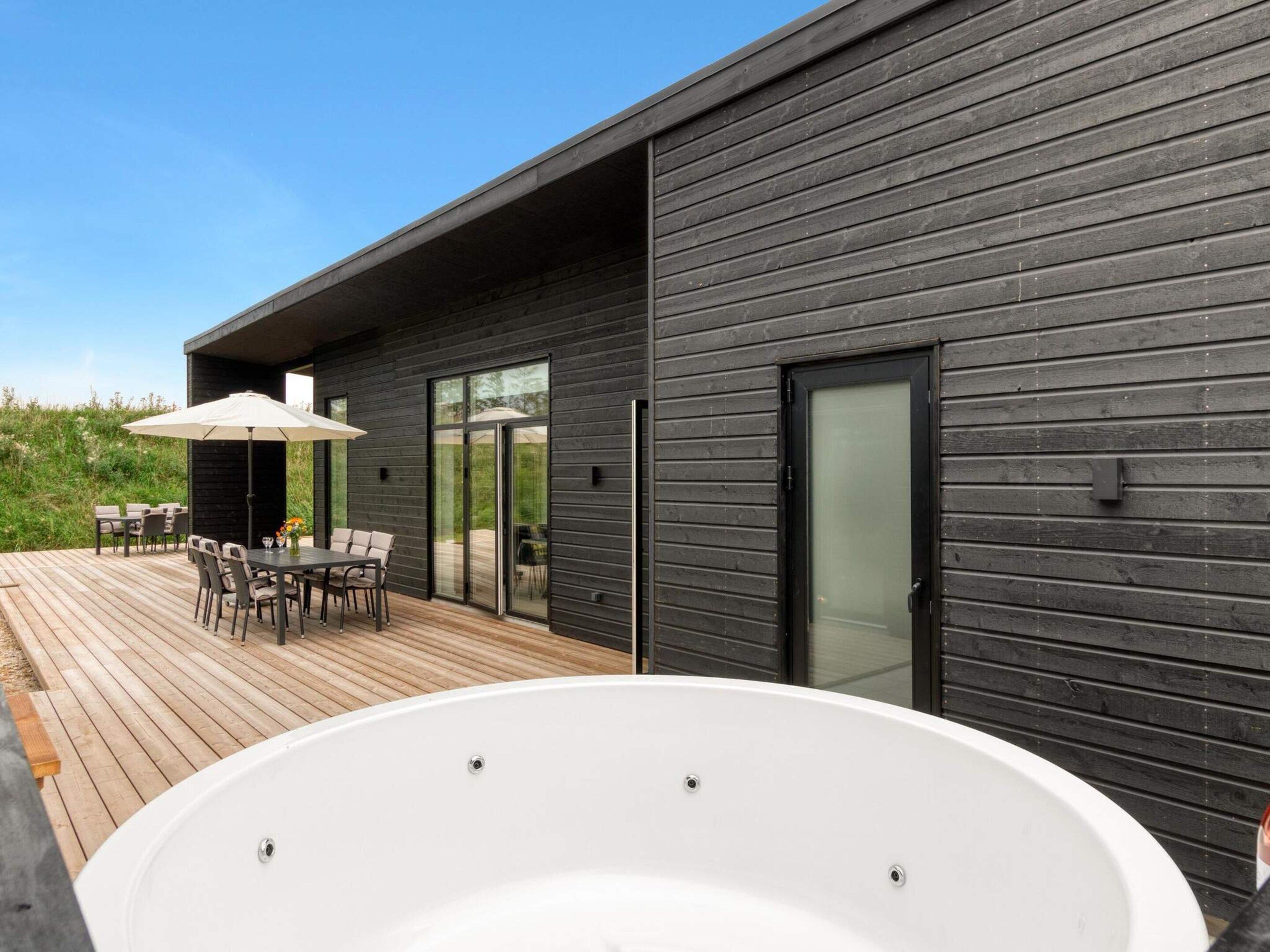 A modern outdoor deck with a hot tub, wooden furniture, and a glass door leading to an outdoor area.