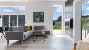 A modern living room with a grey sectional sofa, a coffee table, and a large window offering a view of the outdoors. - 5 Star Holiday Home in Lokken (Lokken)