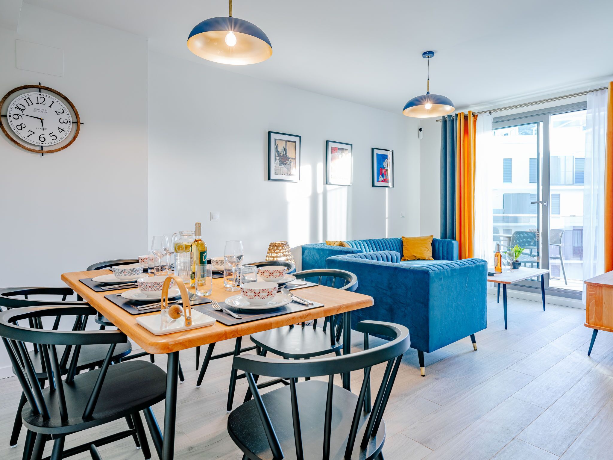 Een moderne woonkamer met een eethoek, met een blauwe bank, een houten eettafel met stoelen, en een groot raam met gordijnen.