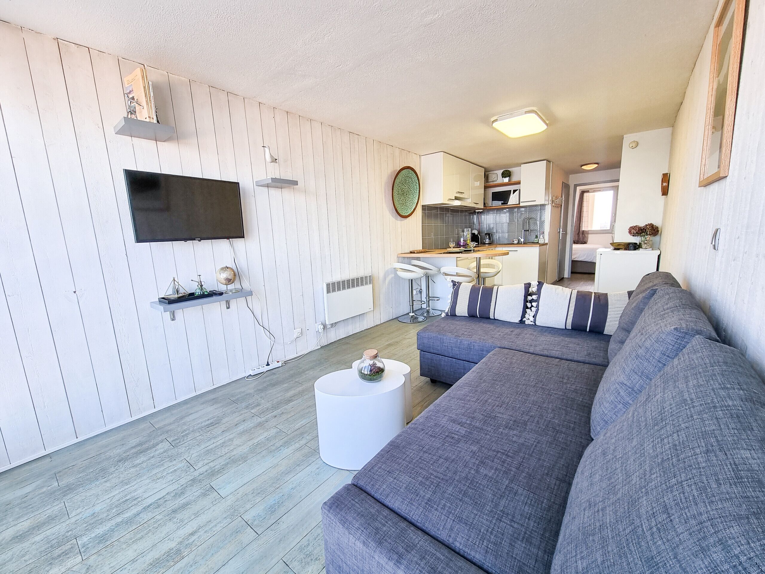 Een moderne woonkamer met een grijze bank, een flatscreen televisie en een keukenhoek met een eettafel en barstoelen.