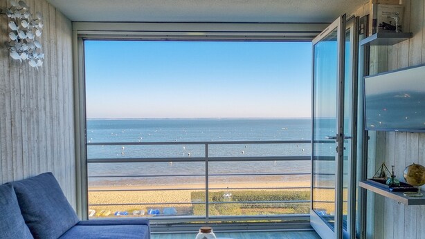 A modern room with a large window overlooking a beach, a sofa, and a small table with a decorative item. - [Vue Imprenable] Appartement Rénové Front de Mer (Arcachon)