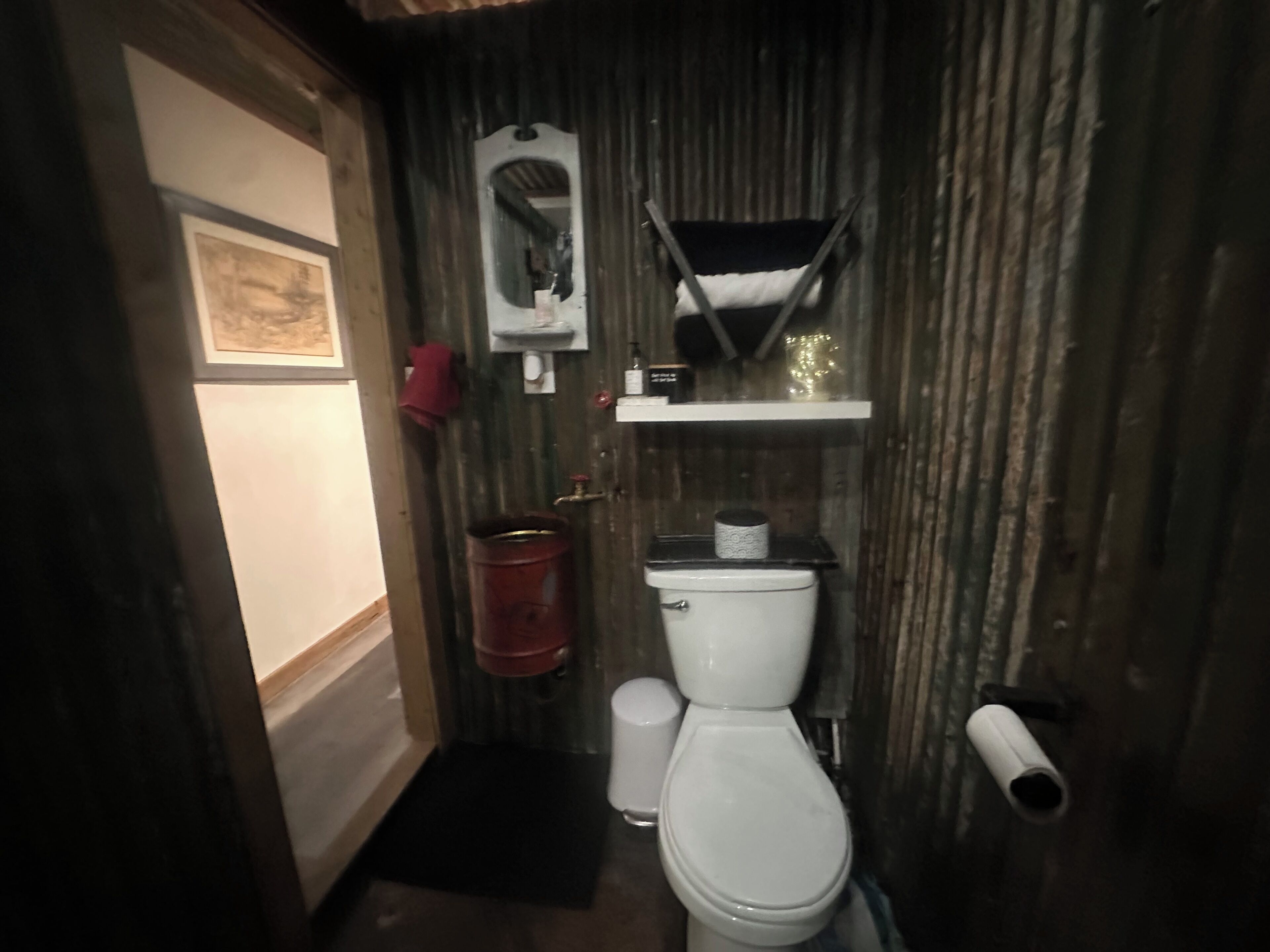 A compact bathroom with a white toilet, a red bucket, and a shelf with toiletries.