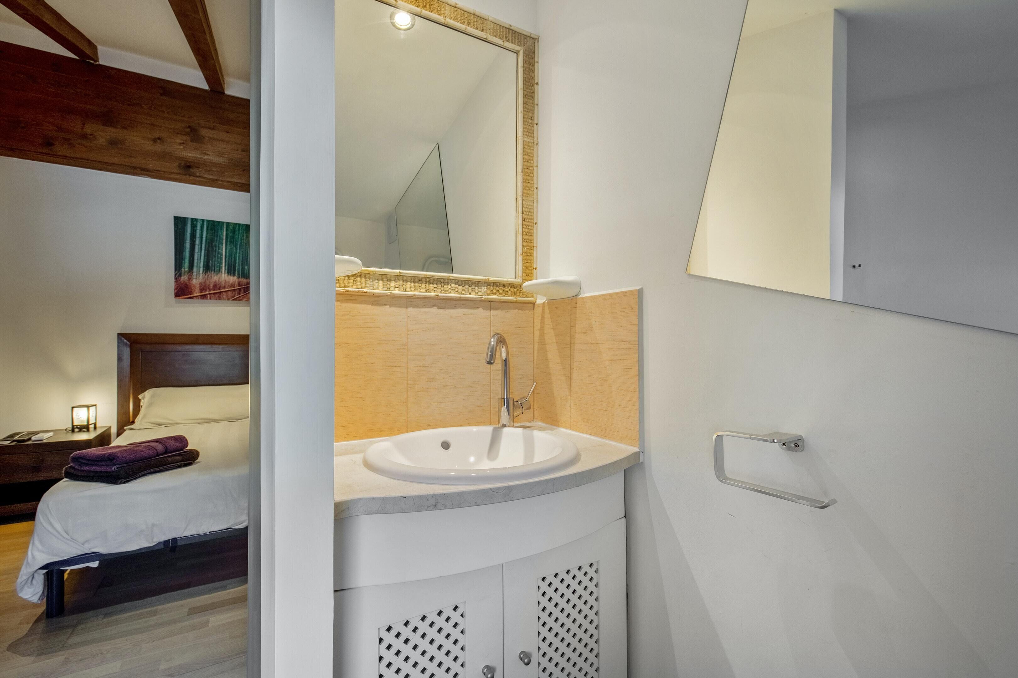 A modern bathroom with a white sink, wooden beams, a mirror, and a bed with a nightstand.