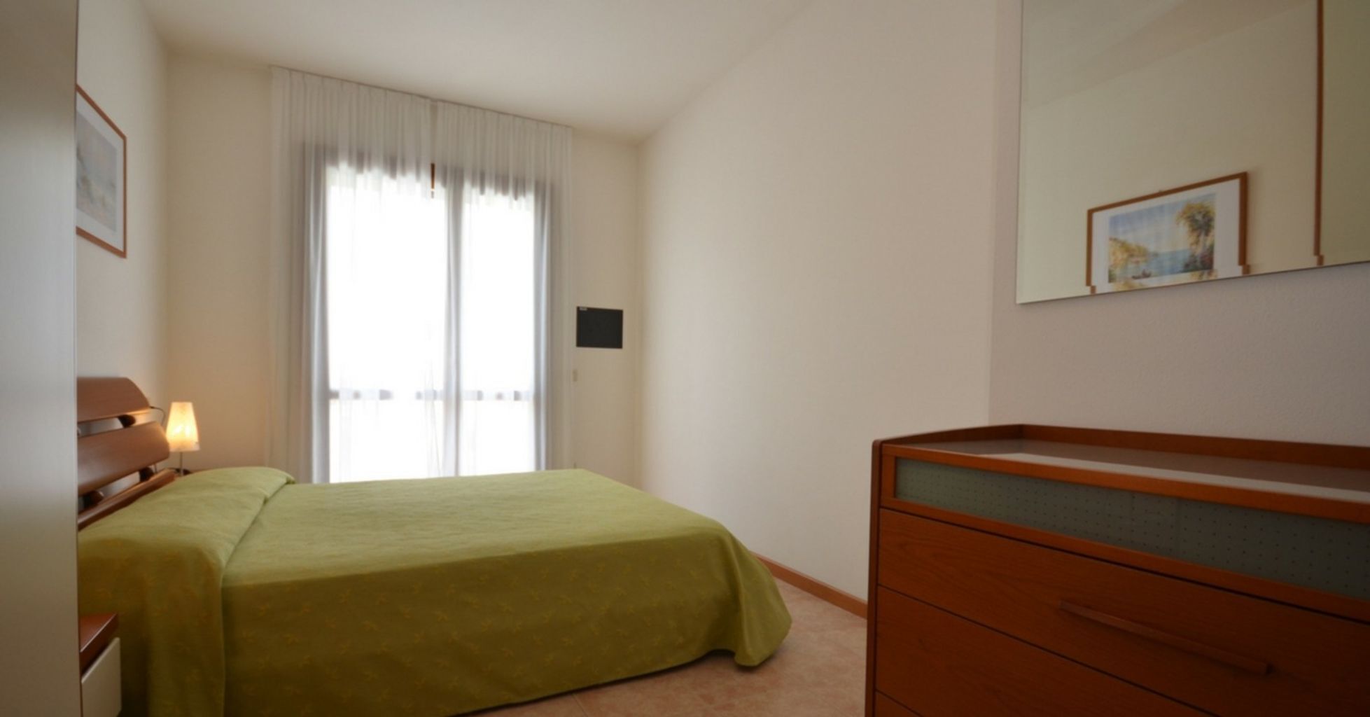 Une chambre à coucher comprenant un lit, une commode, une lampe, une fenêtre avec des rideaux et un tableau accroché au mur.