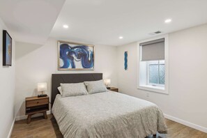 Ein Schlafzimmer mit Bett, Nachttischen, einem Gemälde, einem Fenster mit Jalousie und einer Deckenleuchte.