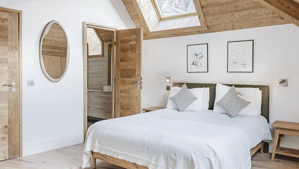 Une chambre avec un plafond en pente, des poutres en bois, un puits de lumière, un lit avec une literie blanche, deux tableaux encadrés au mur et une porte en bois.