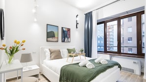 Quarto moderno com uma cama grande, mesa de cabeceira com abajur, pequena mesa com planta e janela com cortinas.