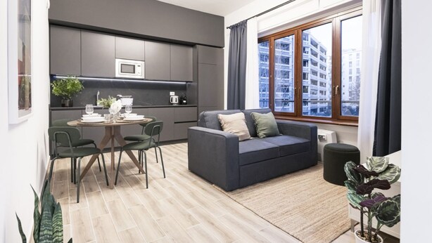 Uma sala de estar moderna com um sofá cinzento, uma mesa de jantar com cadeiras, uma área de cozinha com micro-ondas e uma grande janela com vista para edifícios.